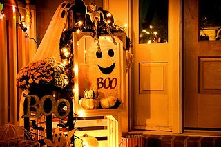 A porch decorated with Halloween decor — ghosts, skeletons, pumpkins, orange and yellow flowers, signs that say “Boo,” and fairy lights.