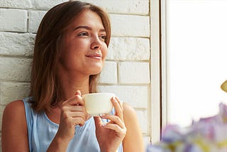 A smiling woman lying against a white wall drinking from a mug