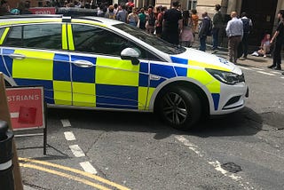 A police car, with a yellow-and-blue Battenberg pattern design, is closing off Clare Street in Bristol city centre to protect Dr Who filming. A crowd watches the filming taking place.