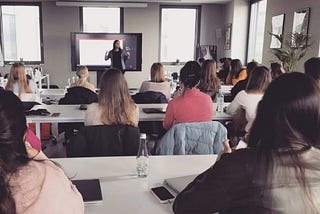 A young woman giving a presentation for a group of a dozen or so people in the room.