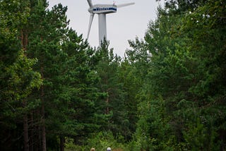 Två barn går längs en skogsväg. Över träden i horisonten tornar ett vindkraftverk upp sig.