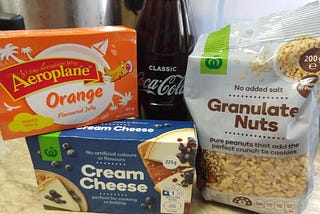 Photograph displaying packages of Aeroplane Jelly, Cream Cheese, Granulated Nuts and a bottle of Coca-Cola all together.