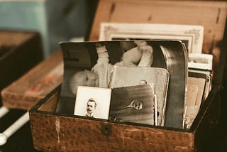 The image, washed out, is of a small wooden box perched atop a table. The box is open and inside rest old photos of people long since deceased that tell the story of someone’s life.