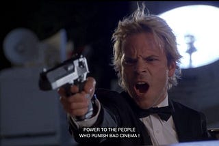 A man with bleached blond hair in a tuxedo points a gun in a studio. He shouts, ‘Power to the people who punish bad cinema!’