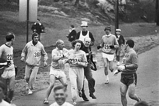 Black and white photo. Kathrine Switzer became the first woman to run the Boston Marathon. Here she is pictured making history while being attacked by the race organizer.