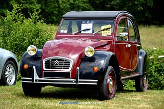 La imagen muestra un Citroën 2CV clásico, un modelo vintage conocido por su diseño distintivo y su importancia histórica en el mundo automotriz. El coche tiene un esquema de colores de dos tonos con una carrocería marrón y guardabarros negros. En la parte frontal del coche, se pueden ver los faros redondos, una parrilla cromada con el logotipo de Citroën y un parachoques negro con detalles cromados. El coche está estacionado en una zona de césped con algo de vegetación en el fondo. También hay algunos papeles o carteles en el parabrisas, posiblemente indicando que el coche es parte de una exhibición o muestra de autos. En la parte inferior de la imagen se puede leer el sitio web "lautomobileancienne.com".