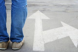 Arrow on pavement pointing straight ahead, with another pointing to the side. Feet standing at that point—indicating making a choice.