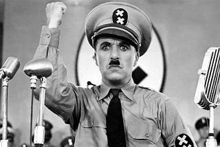 In this film still from The Great Dictator Charlie Chaplin stands dressed in a uniform similar to Hitler , arm raised in a fist pump as a parody of the Nazi leader.
