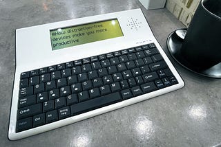 A portable distraction-free writing device with an electronic screen is displayed on a table next to a black coffee cup