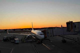 Commercial airplane parked at airport gate during sunset, connected to a jet bridge with ground crew and service vehicles nearby. Warm orange and yellow hues fill the sky, highlighting the serene yet active atmosphere of air travel operations.