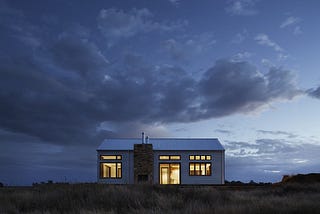 Small home, lights on inside, in the middle of a field, against the fading light in the evening sky.