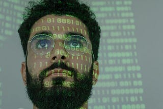 Man with beard and glasses looking up at a reflection of computer code of ones and zeros. Image courtesy of Cottonbro Studios at Pexels
