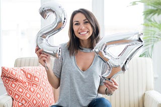 Brunette woman sitting on a white couch with silver “24” balloons for her birthday