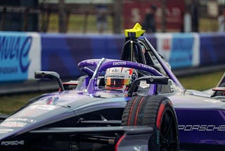 Un coche de carreras de Fórmula E, un Porsche de color morado con detalles en negro y rojo, circula por la pista. El piloto, visible en el coche, lleva un casco con el logotipo de Porsche y otros patrocinadores como TAG Heuer y Loctite. De fondo, una valla publicitaria exhibe el texto 'Formula E World Championship'.