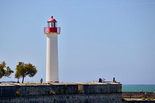 A white lighthouse with a red top stands on a stone embankment in St Martin de Ré, with a few trees nearby and the ocean visible in the background.