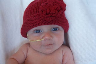 Baby (newborn) Stella with red knit hat, big blue eyes, yellow nasogastric tube smiling angelically at the camera