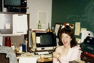 Bitsy as a young software engineer, circa 1984, with vending machine coffee and a pack of Lorna Doone cookies, goofing around in her office at IBM with a microfiche and a stuffed moose on her head.