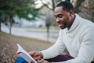 3 Ways to Find Your Work-Life Balance This Year image shows a man in a white sweater and pants sitting by a tree reading with a lake in the background.