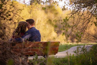 Two people are sitting on a bench on a summer or fall day. They’re smiling and their foreheads are touching.