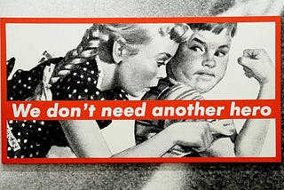 Barbara Kruger’s large black and white illustration of two kids in 5’s advertising style looking a their arm muscles — with red text banner splashed across reading: We don’t need another hero.