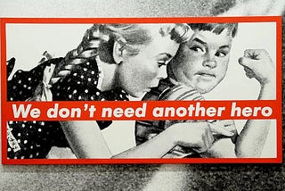 Barbara Kruger’s large black and white illustration of two kids in 5’s advertising style looking a their arm muscles — with red text banner splashed across reading: We don’t need another hero.