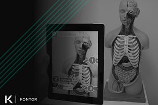 Person holding a tablet pointed at a medical training statue of a human with their internal organs expose. On the tablet, an augmented reality (AR) application is showing organ names in relation to the training statue. On the left side is the Kontor logo and a green Kontor watermark.