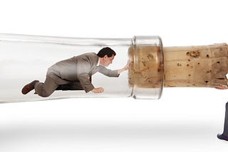 A man is trapped in the neck of a giant corked bottle laying on it’s side. Another man on the outside is pulling on the cork trying to open the bottle.