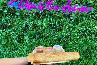 A picture of a plate holding a dosa in front of a backdrop of green plant. Pink neon letters spell out “Dosa is the Answer.”
