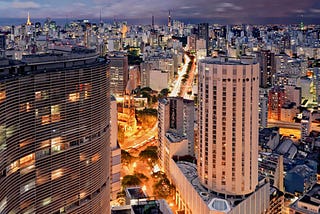 Foto do centro de São Paulo à noite, feita do céu. À esquerda, vê-se parte do edifício Copan, e, à direita dele, um prédio quase tão alto e circular. Ao fundo, veem-se mais e mais prédios, fazendo com que a paisagem seja só concreto e luzes artificiais, e o céu, roxo, como apenas uma faixa fina no topo da foto.
