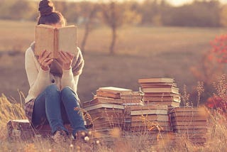 A lady reading next to a pile of books in the middle of a meadow.