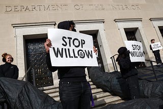a person standing in all black at the department of the interior holding a sign that says “stop willow”