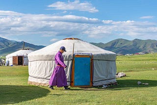 A Mongolian nomad dressed in traditional garb walks outside his ger located on the steeps.