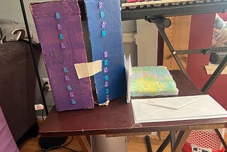 Cardboard box painted purple and blue with two vertical lines of plastic bears glued to the front. A piece of tape is holding the box closed. A fuzzy notebook and a pile of envelopes are on the tray next to the box.