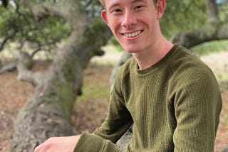 Lucas Morthole smiles while sitting on a low branch of an oak tree.
