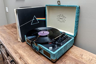 Photo of a record player set in a blue case, with a Ray Charles album ready to play, and Pink Floyd’s The Wall album propped up against it, along with a few other albums, sitting on a wooden table.