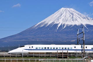 One of the newest Shinkansen series against the background of a wide-reaching Japanese mountain. The train is blue and white, with a head that appears to be in in a 45 degree angle downwards.