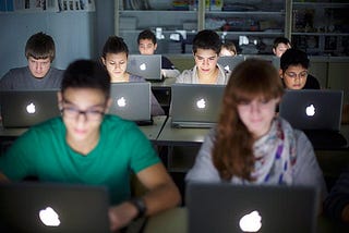 A photograph of nine students sitting in a dark classroom, all working on Apple laptops.