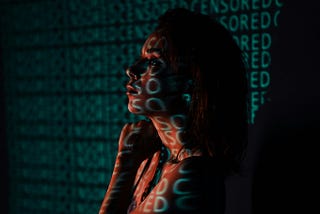 A woman stands in profile with the word “CENSORED” projected repeatedly across her face and body in teal light, suggesting themes of digital suppression and identity erasure.
