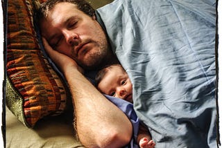 Man sleeping with a baby peeking out beneath the sheets.