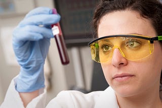 A woman in a white lab coat is wearing protective goggles and gloves, holding a test tube of blood up to the light.