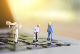 3 figurines of male construction workers wearing yellow hats, standing on a black computer keyboard