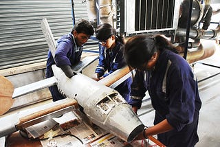 The image depicts three individuals in work uniforms actively engaged in the hands-on maintenance and repair of an aircraft model, demonstrating practical training in aircraft maintenance engineering.