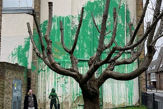 The mural features a mass of green paint splashed across the previously dilapidated wall behind a leafless tree, giving the appearance of the tree’s foliage. A person holding what seems to be a pressure hose has been stenciled alongside it. The artwork has just officially been confirmed as a Banksy; after many had already speculated as such as the mural resembled the style of the street-artist’s previous work.