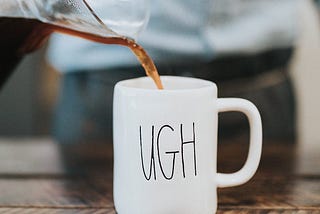 A cup with “ugh” written on it is placed on a hard surface and coffee is being poured from a jug into it.