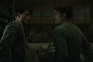 Robert Pattinson as both Mickey 17 and 18, sitting on a bed, looking at each other.