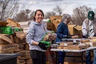 Color photo of food distribution volunteers preparing to share food with the community. A volunteer weraing a gray hoodie and wearing black gloives is holding a large sack of potatoes and smailing.