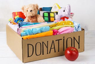 Box with Donation written across the front, filled with clothes, toys, and stuffed animals. Apple in front of box
