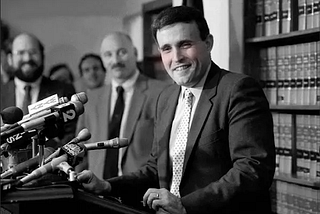 Black and white photo of Rudy Giuliani at a press conference in the 1987. Altered so he appears to be blushing.