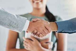 Two male hands shaking with a woman smiling in the background