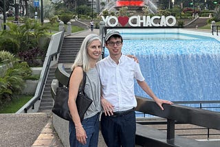 Woman and young man smiling at camera; stading in a plaza with a waterfall behind them